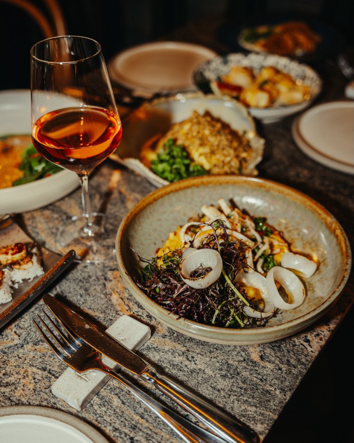 A table in service on dark granite: a glass of orange wine, a bowl of squid and small plates
